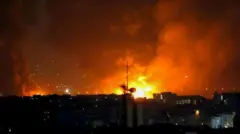 暗い建物群の向こうでオレンジ色の大きな火の手が複数上がり、煙が夜空に立ち上っている 