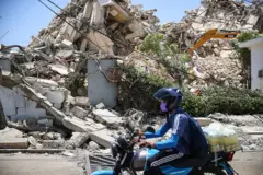 Ağzına maske takmış bir motosiklet sürücüsü, yıkılmış bir binanın önünden geçiyor, motosikletinin arkasında meyve suyu şişeleri taşıyor. 