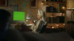 Mulher de roupão, toalha na cabeça e fone de ouvido, sentada em frente ao computador, olhando para o telefone celular, à noite