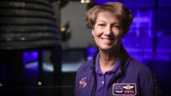 Eileen Collins usa uma jaqueta azul com um emblema da Nasa de um lado e um patch do ônibus espacial com seu nome do outro. Ela tem cabelo castanho curto e olhos castanhos. Ela está sorrindo para a câmera. Está na galeria espacial do Museu de Ciência de Londres e, ao fundo, desfocado, há um motor de foguete.