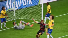 A imagem mostra um lance dentro da área durante uma partida entre Brasil e Alemanha. Enquanto um jogador alemão comemora de braços erguidos, jogadores brasileiros demonstram frustração ao redor da pequena área, com o goleiro caído no gramado segurando a bola e defensores observando a cena próximos à linha do gol.