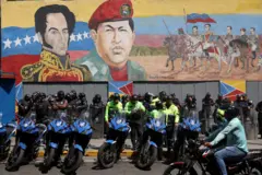 Fileiras de policiais de pé atrás de motos, usando capacetes em frente a um enorme mural pintado de Simón Bolívar e Hugo Chávez