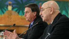 Osmar Terra fala ao microfone, com Jair Bolsonaro no plano de fundo. Ambos estão sentados diante de mesa em sala
