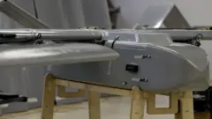 Um drone cinza, parecido com um pequeno avião, repousa sobre uma estrutura de madeira em uma sala comum.