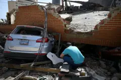 Un hombre acuclillado revisa el suelo alrededor de un auto aplastado por un pedazo de edificio que le cayó encima en los terremotos.