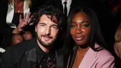 Andrea Preti wear black suit e get curly hair and e smile to di camera. Venus William wear pink suit wit black long hair wey dey fall for her shoulders. She smile to di camera too.