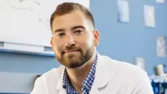 Ben Rein, com cabelo castanho claro curto e barba castanha, fotografado em ambiente interno, usando jaleco branco de laboratório e camisa xadrez azul e branca