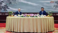 Los presidentes Putin y Xi miran a la cámara sentados en una mesa redonda con un mantel dorado y flores encima; en el fondo se ve una pintura china.