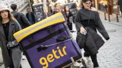 Getir logolu bir araç park etmiş etrafından iki kadın yürüyor. 