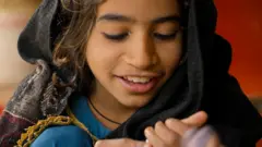 Una niña con un vestido azul sonríe, con la mirada fija en sus manos. Lleva un pañuelo oscuro con un borde estampado sobre la cabeza. 