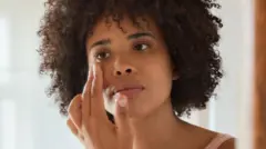 A young woman with dark hair looks in the mirror, touching the skin close to eye.