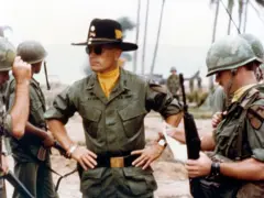 Robert Duvall interpretando o tenente-coronel Kilgore, rodeado por soldados. Duvall está no centro, usando um grande chapéu preto, óculos escuros e uma bandana amarela.