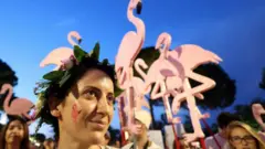 A young woman with a wreath on her head and a flamingo painted on her cheek is seen in front of flamingo-shaped placards at a protest in Albania