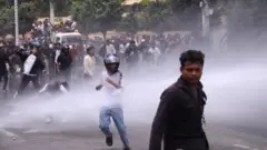 Un hombre con casco de motocicleta, pantalones azules y camiseta blanca corre bajo un chorro de gas o agua mientras la multitud lo sigue, el 8 de septiembre de 2025. Un hombre levanta el brazo, como si lanzara un objeto. Un hombre en primer plano mira a la cámara.
