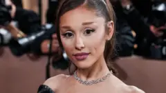 A imagem mostra Ariana Grande posando para fotógrafos em um evento de tapete vermelho. Ela aparece em primeiro plano, olhando diretamente para a câmera, com os cabelos presos em um rabo de cavalo, maquiagem discreta, brincos e um colar de diamantes. Veste um traje preto de ombros à mostra, enquanto, ao fundo, fotógrafos e câmeras desfocados reforçam o ambiente de uma premiação.