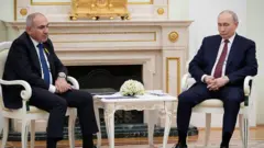 Sol tarafta Nikol Paşinyan, sağ tarafta Vladimir Putin sandalyede oturuyorlar. Ortalarında beyaz bir sehpa bulunuyor. İki lider de önüne bakıyor. 