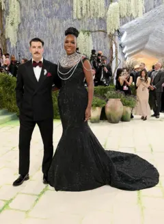 Un hombre y una mujer posan en una alfombra verde durante un evento formal; él viste traje negro con pajarita y ella un vestido largo negro con joyería. Hay fotógrafos y decoración floral de fondo.