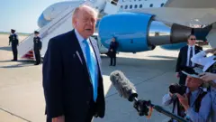 Trump takım elbise giyiyor ve muhabirlerle konuşuyor, arkasında bir uçak park etti.