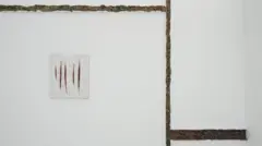 Em uma parede branca minimalista, uma pequena obra de azulejos exibe cortes verticais que lembram feridas abertas, enquanto estruturas orgânicas e escuras percorrem o espaço como fissuras ou cicatrizes.