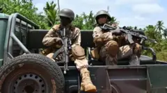 Aworan awon okunrin meji to dihamọra ogun pẹlu ibọn lọwọ, wọn bo oju, wọn jokoo ninu ọkọ ologun.