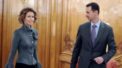 بشار الأسد (يمين) مرتدياً لباساً رسمياً بلون رمادي غامق وبجانبه زوجته أسماء مرتدية قميصاً رمادياً فاتح اللون
