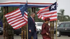 El expresidente estadounidense George W. Bush junto a la ex presidente de Liberia Ellen Johnson Sirleaf junto a las banderas de EE.UU. y de Liberia. La de Liberia tiene menos franjas rojas y blancas que la bandera estadounidense, y lleva una sola estrella blanca sobre el cuadro azul.