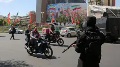 یکی از میدانهای تهران در حالیکه مامور پلیسی که صورت خود را پوشانده و اسلحه در دست دارد ایستاده. عابران و دو موتور سوار در حال گذار از خیابان هستند.