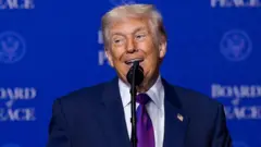 Trump hablando ante un micrófono, con un fondo azul donde se lee “Board of Peace”.