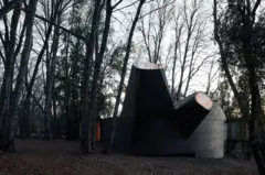 Estructura arquitectónica de formas mayormente rectas en un bosque de árboles altos y desnudos al atardecer.