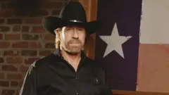 Chuck Norris usa uma camisa preta e um chapéu de caubói enquanto posa para uma foto. Atrás dele, vê-se uma bandeira do Texas e uma parede de tijolos.