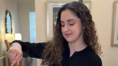 Uma mulher vestida com blusa preta está de pé, inclinada sobre a bancada da cozinha. Ela tem cabelos longos e cacheados, com tons castanhos caindo sobre os ombros, segura uma colher de madeira e parece mexer um alimento.