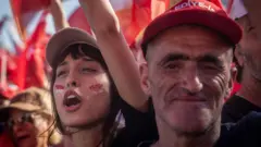 19 Mayıs 2025'te İzmir'de yapılan CHP mitinginde, bir belediye işçisi ile genç bir partili kız yan yana. Kız slogan atıyor, işçiyse fotoğrafçıya bakıyor.