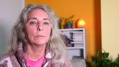 Um retrato fechado, em ambiente interno, de Janet já adulta, com cabelo longo, ondulado e claro. Ela usa uma blusa rosa e um fleece estampado. Ao fundo há um ambiente colorido, com parede amarela, prateleiras, livros, plantas e uma luminária acesa suavemente