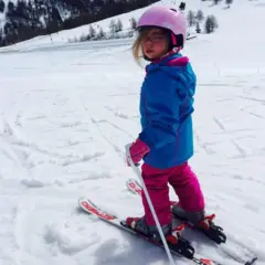 Sophia de niña, sobre esquís en la nieve. Tiene el cabello largo y rubio, lleva gafas y viste un casco rosa, una chaqueta impermeable azul y pantalones de esquí rosas. Sostiene un bastón de esquí y en sus esquís hay inscripciones en rojo. Está mirando a la cámara.