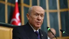 MHP Genel Başkanı Devlet Bahçeli kürsüde konuşma yaparken görülüyor. Arkasında MHP bayrağı ve uzakta bir kişi var. 