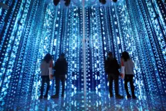 파란색 빛의 디지털 공간에서 밝은 숫자와 기호들이 벽과 바닥을 가득 채우고 있으며 관람객이 미디어 아트를 감상하고 있다