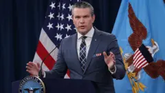Pete Hegseth, cercado por bandeiras, em uma coletiva de imprensa. Ele usa um lenço com as cores dos EUA no bolso do paletó