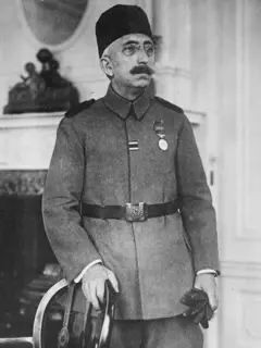 Retrato del sultán Mehmed VI. Tiene puestos lentes y un tarboosh en la cabeza. Viste un uniforme, con un cinturón negro y le cuelga una medalla en el lado izquierdo de su pecho. Con una mano sostiene unos guantes negros y con la otra tiene agarrada una silla.