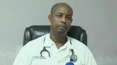 Dr Emmanuel Semwaga yicaye yegamye mu ntebe y'umusego y'umukara, yambaye ishati y'umweru n'itaburiye y'umweru yanditseho izina rye. Mu ijosi yiteye igikoresho cyo kwa muganga cyo gusuzumisha. 