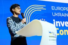 Еврокомиссар по вопросам расширения Марта Кос в синем платье с клетчатыми рукавами с микрофоном в руках выступает с белой трибуны на фоне бело-желто-голубого стенда, на нем справа части слов "Европа" и "Украина"