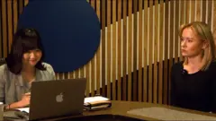 Uma mulher asiática olhando para um computador. Do lado dela há uma outra mulher, sentada.