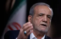 بزشكيان يظهر من الأعلى بشعره الرمادي وبشرته الحنطية، مرتدياً قميصاً أبيض تظهر ياقته من تحت سترة بدلة رسمية بلون أسود. يحرك يده وفيها خاتم فضي كبير، وخلفه علم إيران.