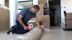 Um homem de camisa polo cinza e calças escuras está ajoelhado no chão, fechando com fita adesiva um grande objeto embrulhado em plástico bolha. Atrás dele, há caixas de papelão e outro homem observando.