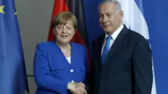 Angela Merkel e Benjamin Netanyahu posam se cumprimentando, em frente às bandeiras dos seus países e da União Europeia
