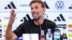 Jurgen Klopp akoze ikimenyetso n’ikiganza, mu kiganiro n’abanyamakuru ku itariki ya 24 Nyakanga (7) mu 2026. Yambaye umupira w’umukara w’amaboko magufi, arimo kuvugira mu ndangururamajwi, imbere ye hari ibyo kunywa byoroheje biri mu macupa, birimo n’amazi.