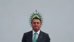 Bolsonaro em pé em frente a painel com símbolo brasileiro