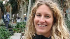 Natalie Mueller, de cabelo loiro comprido, sorrindo para a câmera em rua arborizada