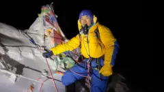Nima Rinji Sherpa, in bright yellow and blue climbing gear, stands in front of an icy peak that is covered in prayer flags