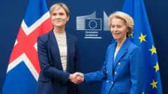 A primeira-ministra islandesa, Kristún Frostadóttir, à esquerda, ao lado da presidente da Comissão Europeia, Ursula von der Leyen. Elas sorriem para as câmeras, com as mãos dadas. Atrás delas, estão as bandeiras da Islândia e da União Europeia.