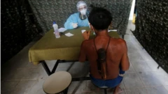 Em tenda de atendimento, homem indígena de costas é atendido, do outro lado da mesa, por profissional de saúde todo coberto por jaleco e face shield